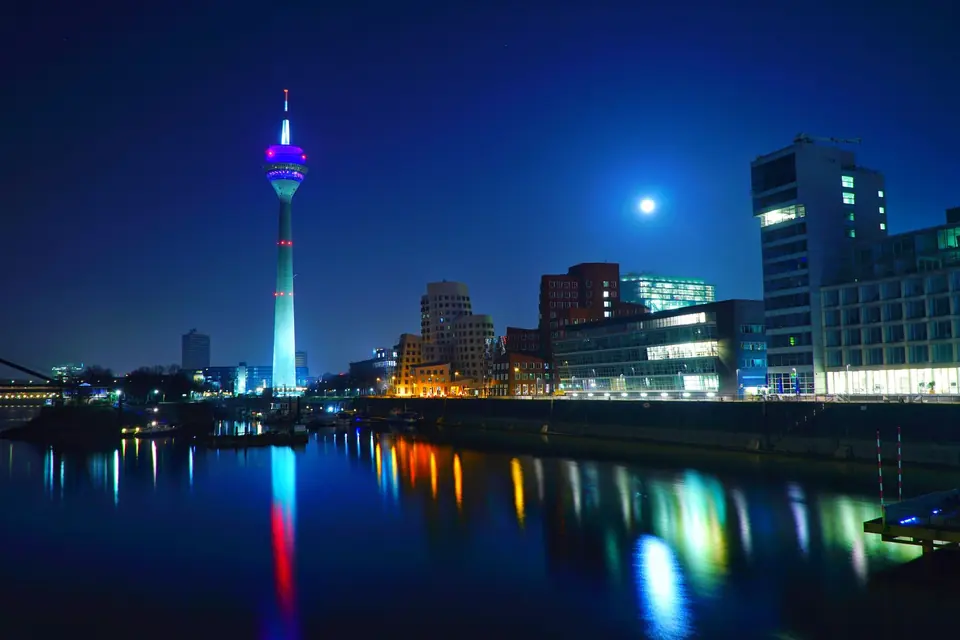 Szenische Nachtansicht der Düsseldorfer Skyline mit dem beleuchteten Rheinturm, der sich im Fluss spiegelt.